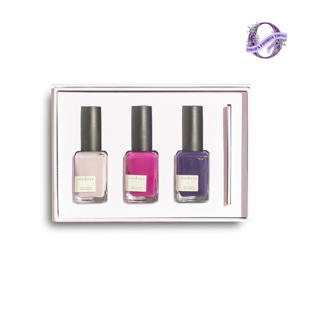 A gift box containing three bottles of nail polish in different shades of purple, with a brush for application, presented against a white background.